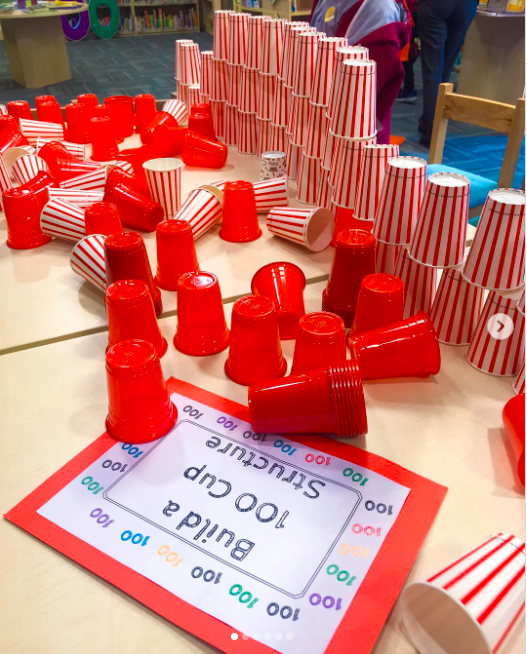 Red plastic cups on a table are set beside a label that says "build a 100 cup structure"