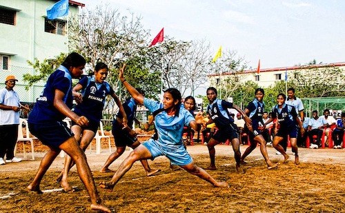 Several older children are engaged in a game on a dirt court. A girl in a light blue shirt is shown with her legs and arms outstretched toward two girls in darker blue shirts. They are playing an international game that can be used as part of American recess games as well.