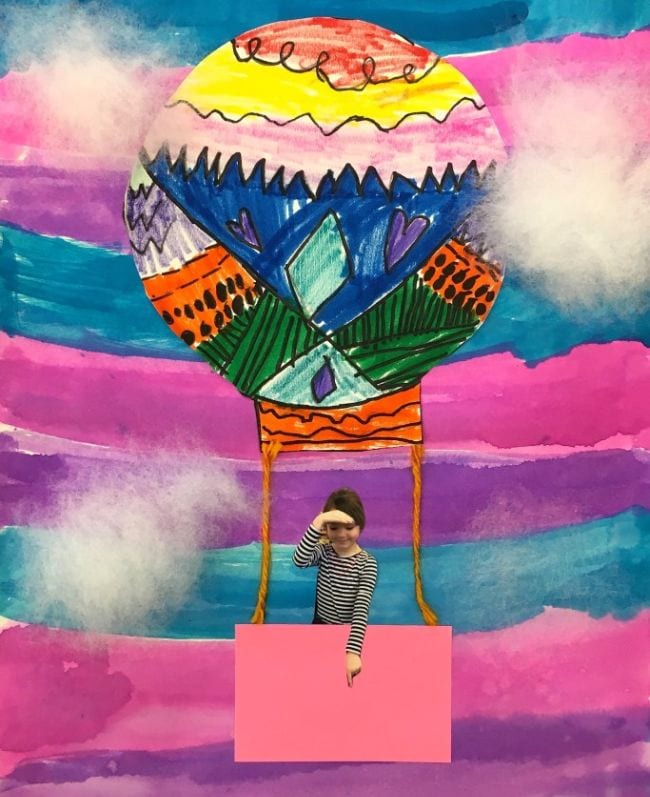 Brightly-patterned paper hot air balloon on a marker-striped background with the photograph of a child in the basket