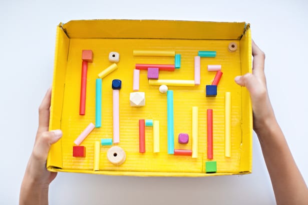 A child's hands are shown holding a yellow box. There are straws and other objects laid out inside to form a maze (earth day crafts)