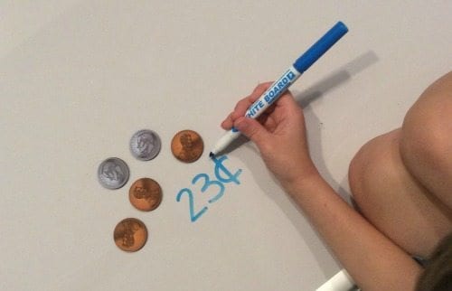 Child writing a dollar amount next to a pile of coin math manipulatives