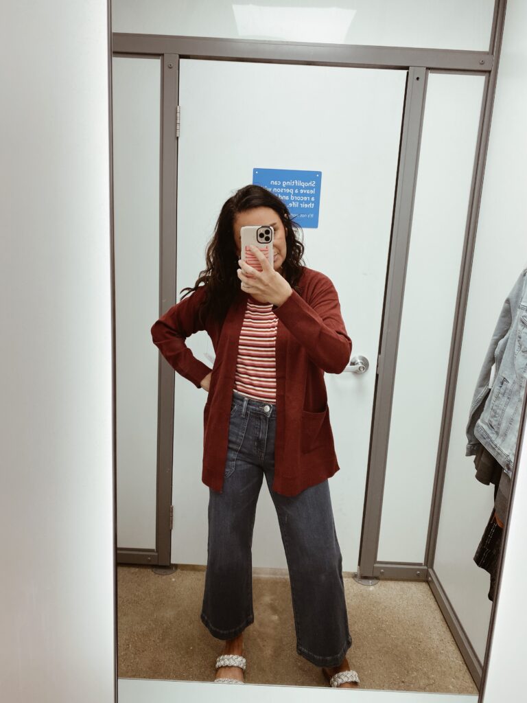 Woman posing in mirror wearing jeans and a cardigan over a striped shirt as an example of cute teacher outfits