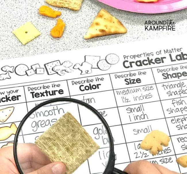 Child's hand holding magnifying glass over a cracker, with worksheet labeled Cracker Lab in the background (2nd Grade Science)