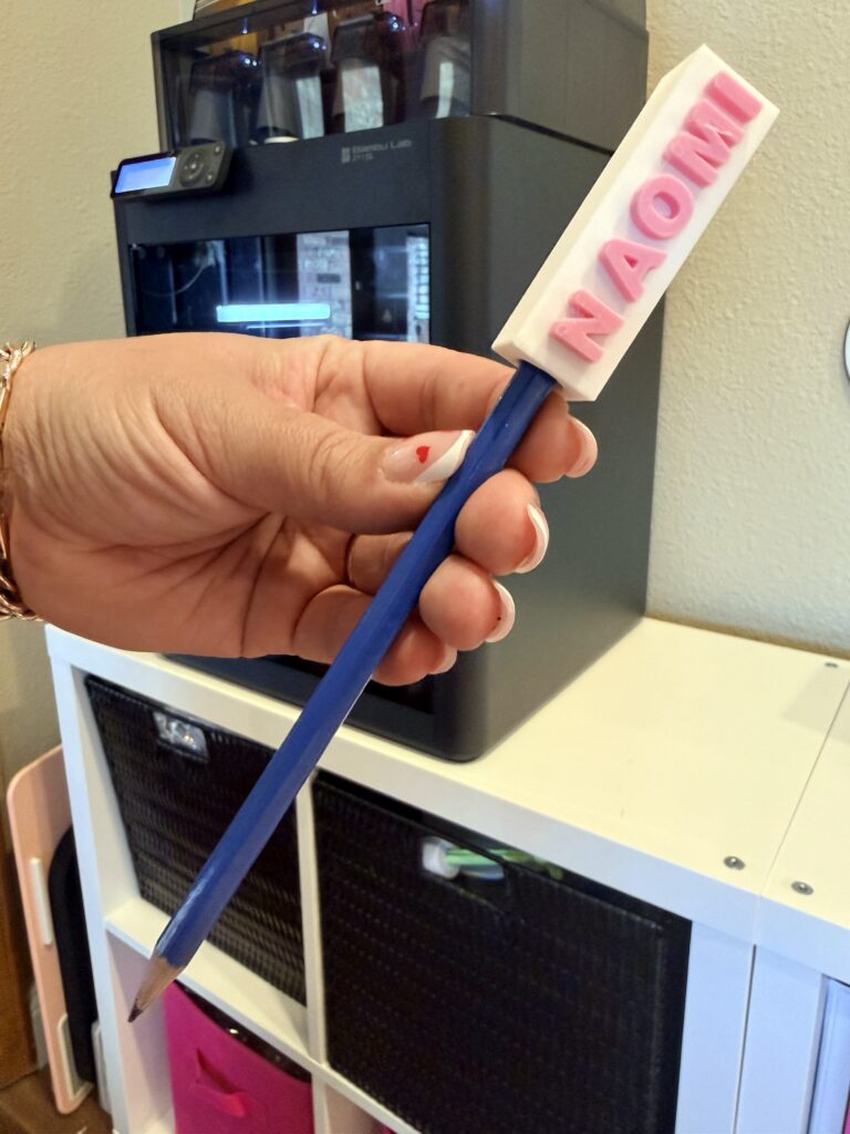 Woman's hand holding a blue pencil with a pink and white 3D printed pencil topper reading Naomi