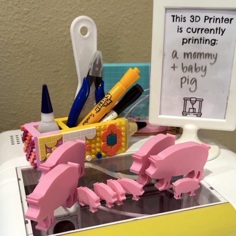 Family of pink 3D printed pigs sitting on top of a 3D printer with a sign indicating what's printing