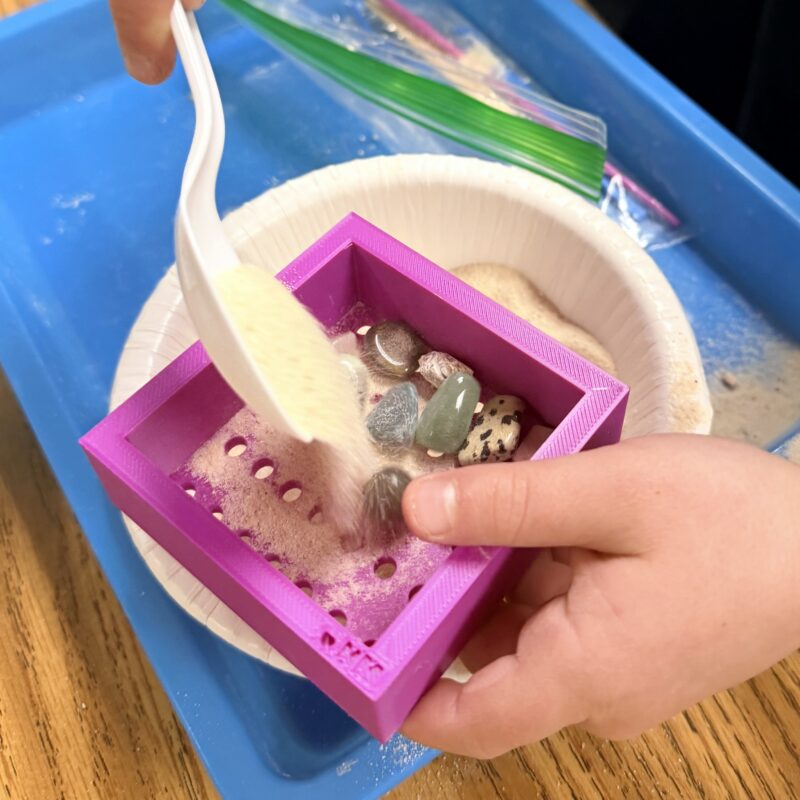 Child using a spoon to pour sand through a 3D printed sifter into a bowl