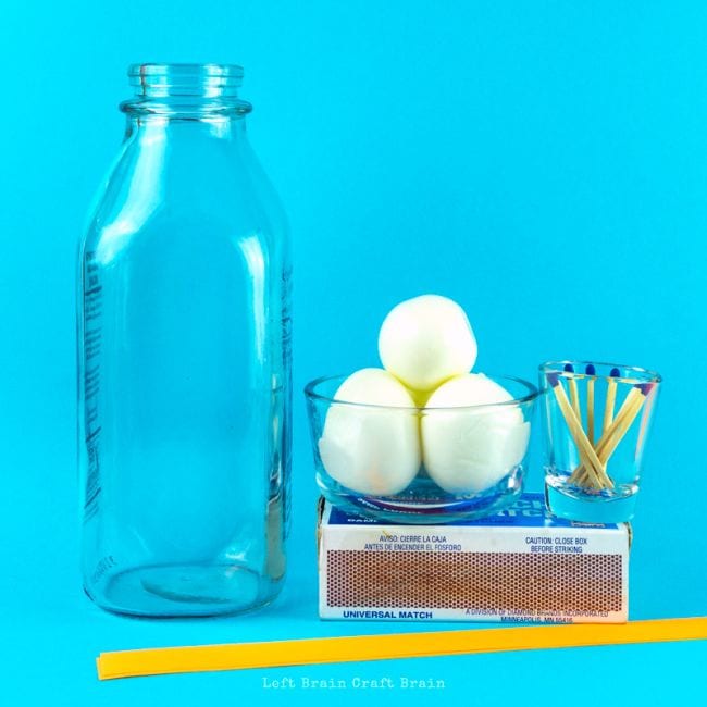 Glass bottle with bowl holding three eggs, small glass with matches sitting on a box of matches, and a yellow plastic straw, against a blue background