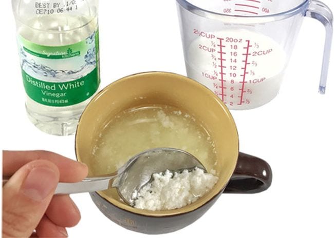 Bottle of vinegar, measuring cup of milk, and a hand using a spoon to scoop plastic bits from a coffee mug 