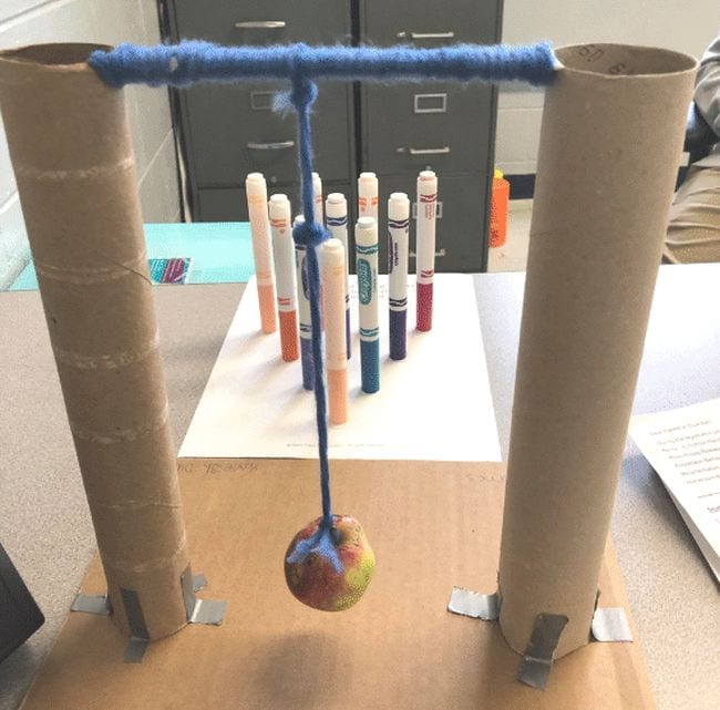 Wrecking ball made from an apple suspended from a rod between two upright paper towel tubes, aimed at an arrangement of markers