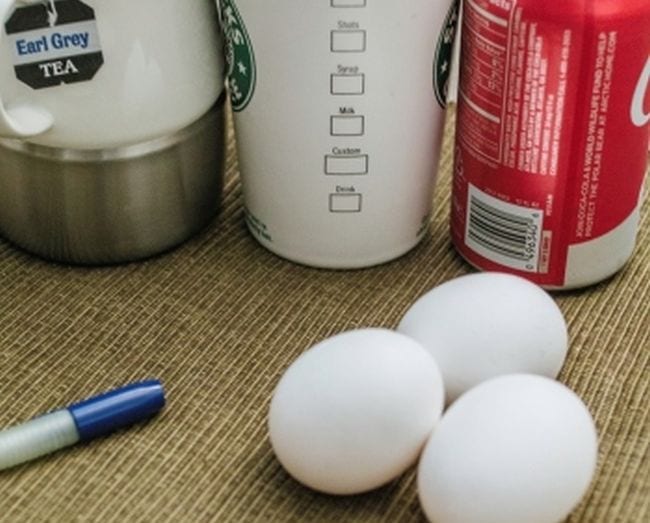 Three eggs next to containers of coffee, tea, and cola, with a permanent marker 