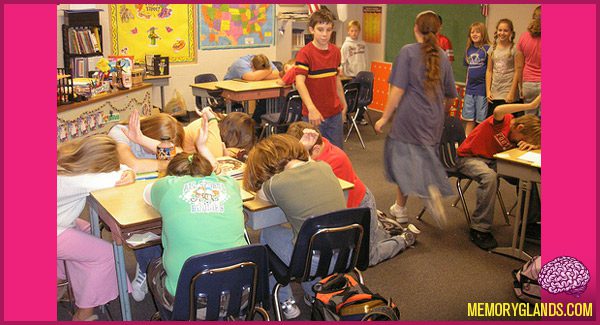 A classroom is shown with several students with their heads down on their desks and their arms extended with thumbs up. Several more students are shown walking around the seated students.