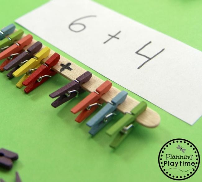 Wood craft stick with colorful clothes pins clipped to it, and a plus sign written in the middle. Above is a card with an addition fact written on it.