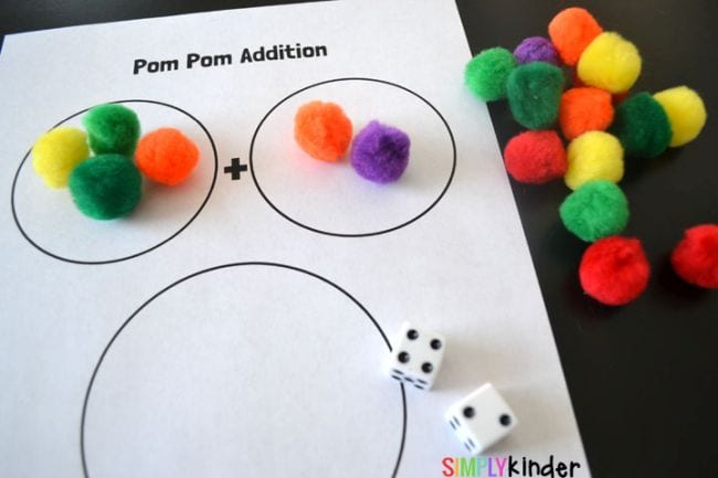 A piece of paper with two small circles and one large one. The small circles each have several pom poms in them. Also shown: two dice and a pile of colorful pom poms.