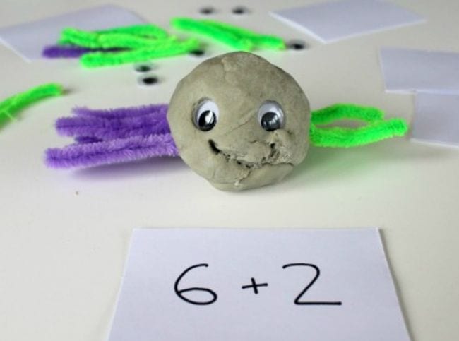 Ball of playdough turned into a slider with wiggly eyes and pipe cleaner legs. One side has purple legs and the other are green. An addition fact is written on a card in front of the spider.