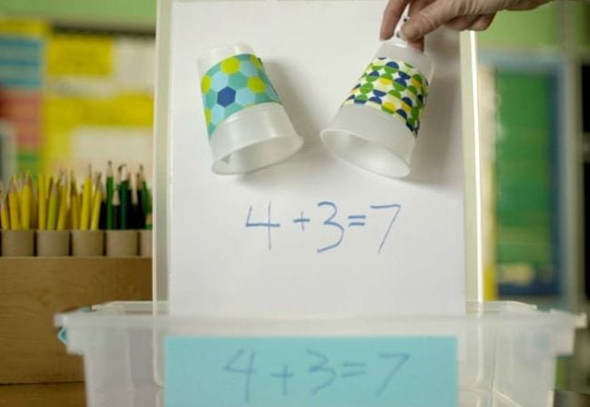 Two upside-down cups with the bottoms cut out mounted to a board. Student is dropping dice through them into a box below, and a math equation is written on the board