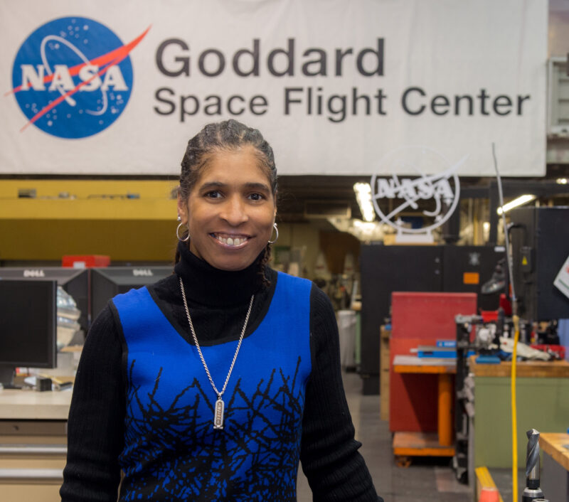 A smiling woman in a blue dress stands in front of a NASA sign.