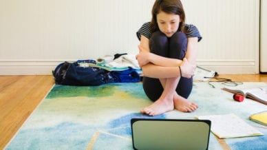 Sad female teenager watching media on laptop hugging herself sitting on the carpet with homework material and clothing around her.