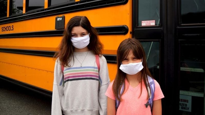 Two young girls standing next to the school bus while wearing white masks.
