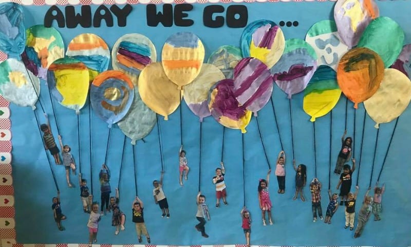 Bulletin board featuring balloons colored by kids, with a cutout photo of a kid hanging from each string (Back to School Bulletin Board Ideas)