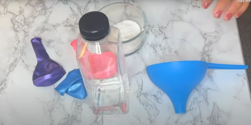 A few deflated balloons, a clear container with clear liquid, a small bowl with baking soda, and a funnel are shown on the table in this step of the baking soda and vinegar balloon experiment.