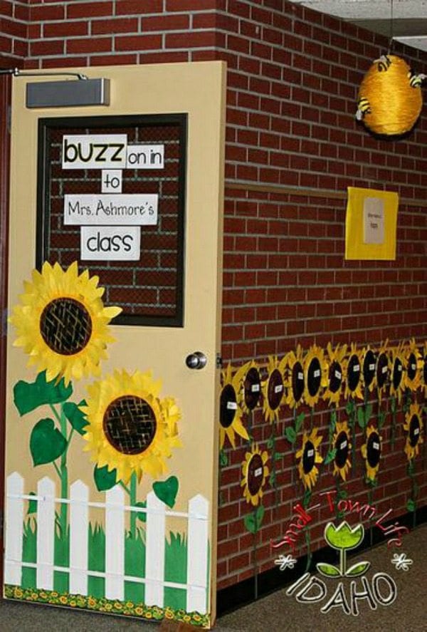 Fall bulletin boards like this classroom door leads ino a hallway. The door reads buzz on in to Mrs. Ashmore's class. There are two large sunflowers on the door and a bunch of smaller sunflowers lining the halls with students' names on them.