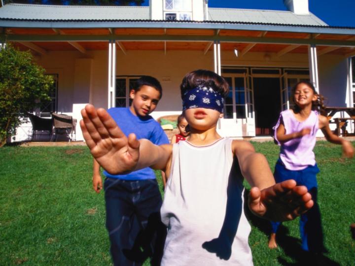 A little boy is shown with a blindfold on and his hands and arms outstretched. Three children are following him from behind.