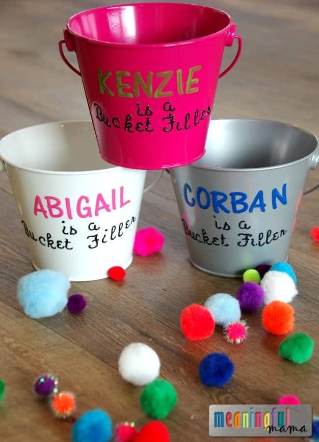 Small metal buckets with text like "Kenzie is a bucket filler" and an assortment of fuzzy pompoms as an example of bucket filler activities