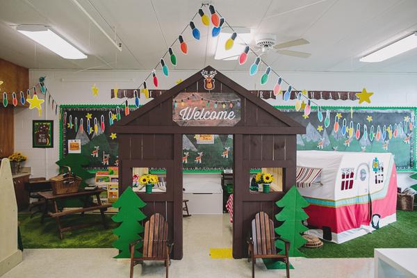 A classroom is setup to look like a campsite. A log cabin entrance is shown with two adirondack chairs. Some pretend trees, a pop up camper, and a picnic table are in the background. 