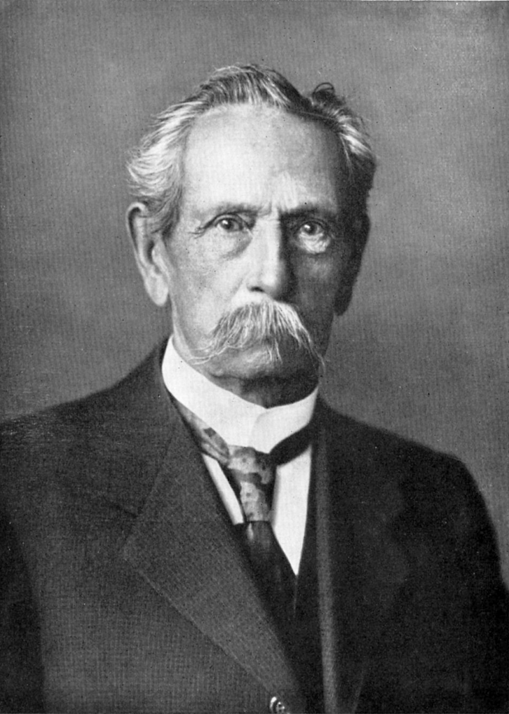 A black and white photograph of an older man with a large moustache is shown from the waist up.