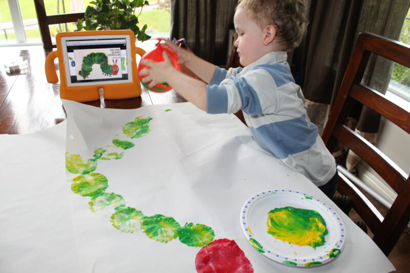 A child holds a balloon over a large white piece of paper that has green and red dots printed on it with paint to look like a caterpillar.