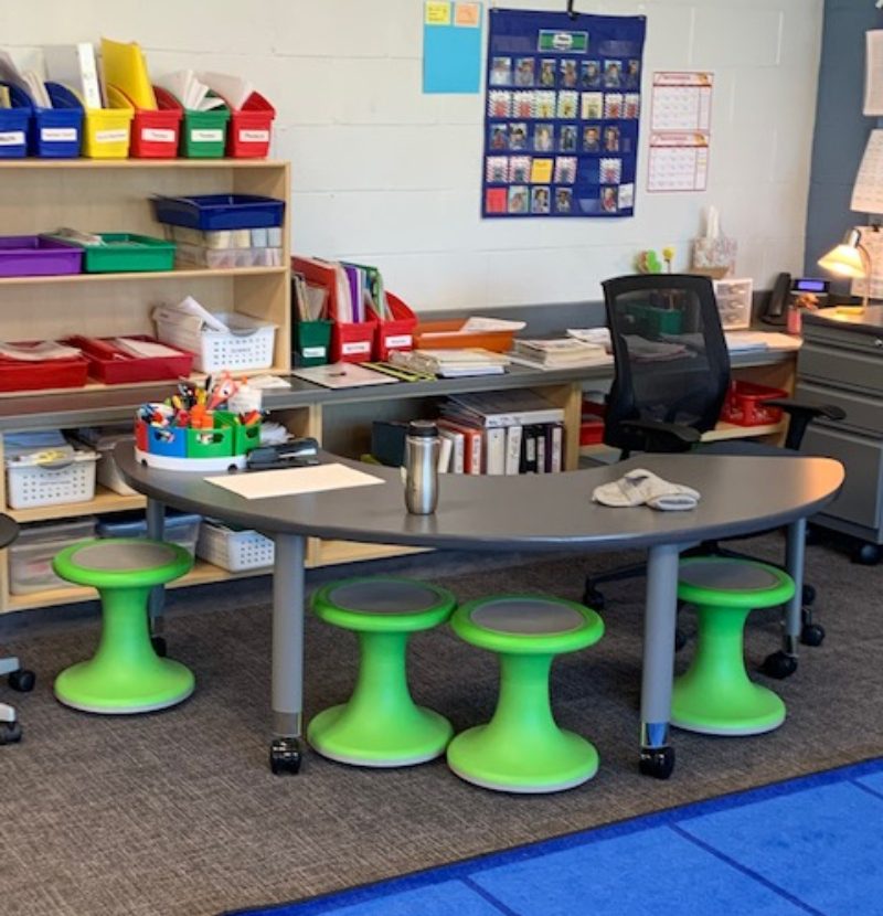 Teacher area in classroom with kidney table and wobbly stools and shelves filled with colorful bins of materials