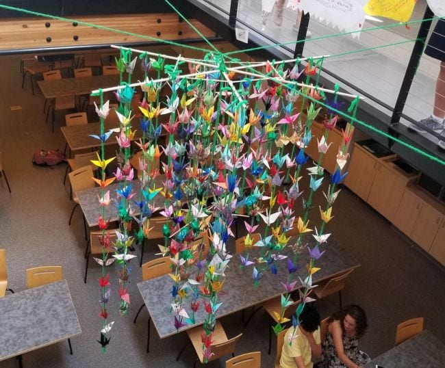 Large mobile made of colorful paper cranes photographed from the top looking down over desks in this collaborative art project.