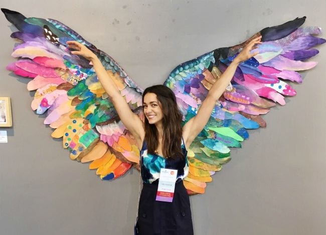 Woman posing in front of large wings made of individually decorated paper feathers