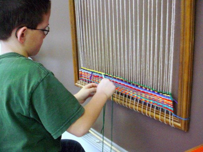 Child weaving yarn on a large loom mounted on a wall 