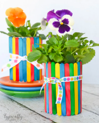 flower pot made from colorful craft sticks glued around a tin can, with colorful flower planted inside, as an example of summer crafts for kids