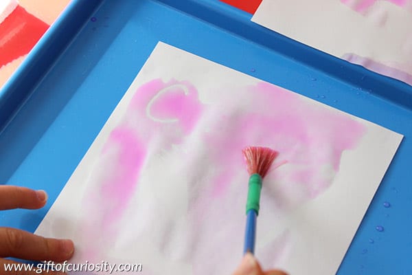 A piece of paper with pink paint and white letters underneath with a paintbrush laying on top