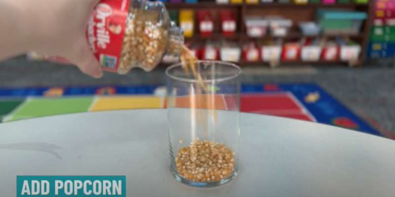 A hand is seen pouring popcorn kernels into a glass jar. Text reads Add popcorn. 