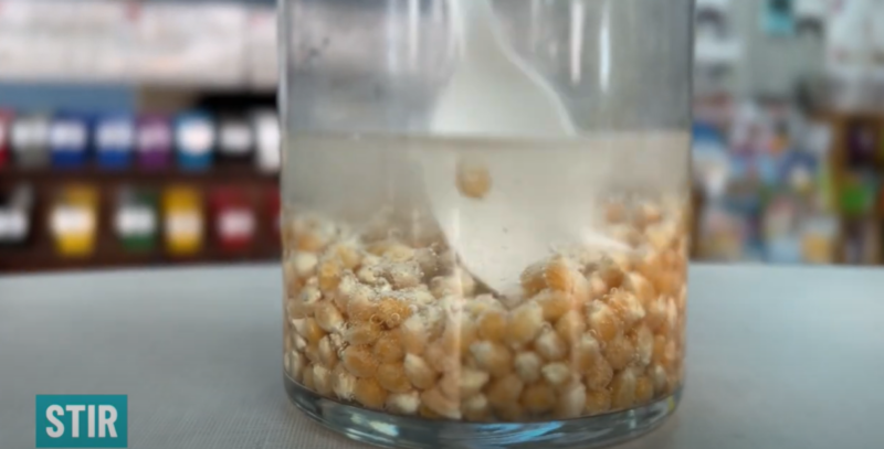 A plastic spoon is seen inserted inside a glass jar containing liquid and popcorn kernels. Text reads Stir.