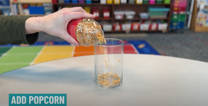 A hand is seen pouring popcorn kernels into a glass jar in this first step in the dancing popcorn experiment. 