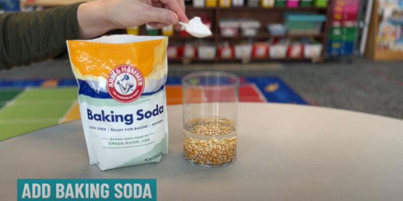 A hand is seen holding a spoon with a pile of baking soda on it. A bag of baking soda is also shown as is a glass jar filled with liquid and popcorn kernels.