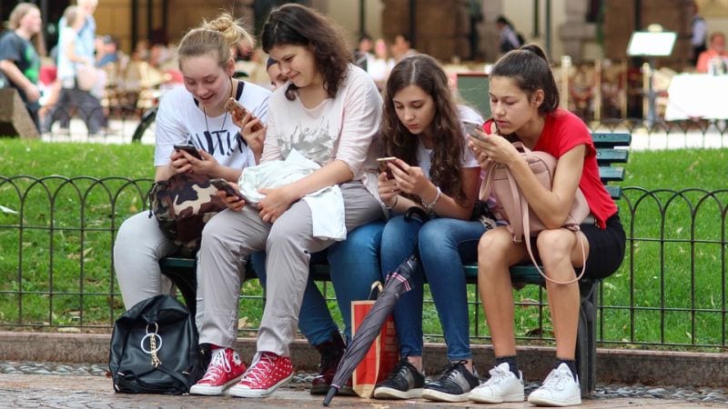 Group of teen girls sitting on a park bench looking at their phones.