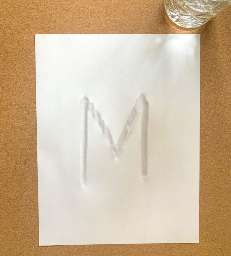 Letter M drawn with water on white paper with a jar of water next to it 