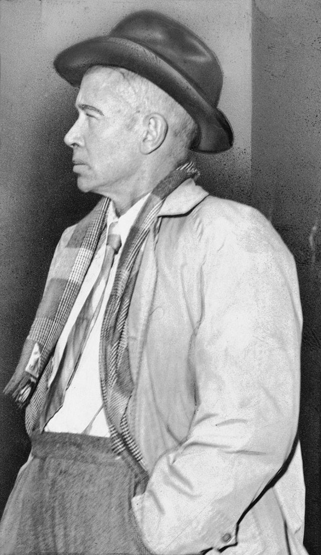 Black and white side view photograph of E. E. Cummings wearing jacket and hat with hands in pockets.