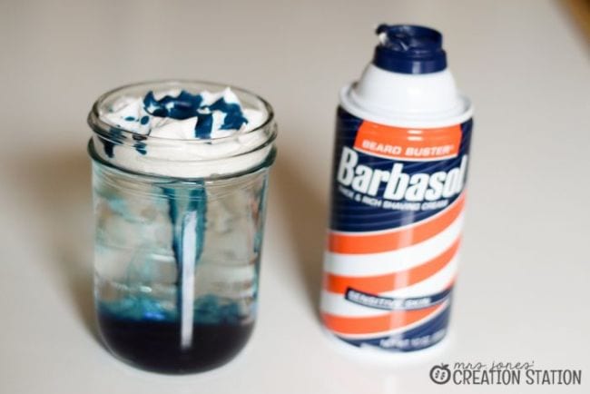 Glass jar of water with shaving cream floating on top, with blue food coloring dripping through, next to a can of shaving cream