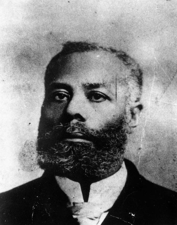 A black and white headshot of a bearded man with a serious expression is shown. 