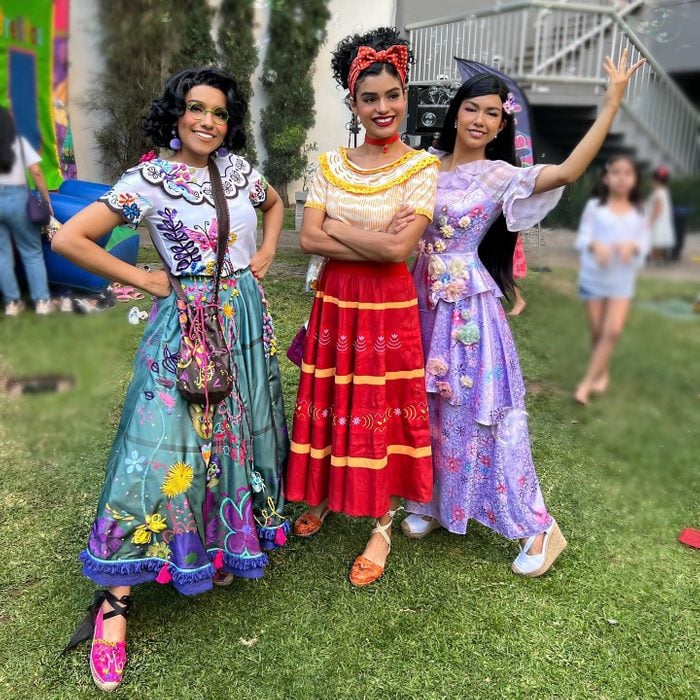 Three women are shown dressed as Mirabel, Dolores, and Isabel from Encanto. The woman on the left has on a white and teal dress, the woman in the middle has on a yellow, white, and red dress, and the woman on the right has on a fancy purple dress.