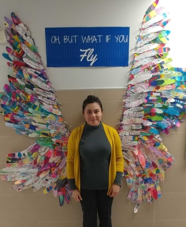 A woman stands in between two wings attached to a wall. The wings are made from feathers that students have colored and written on. Text reads 