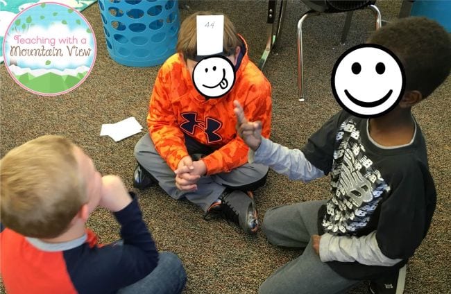 طلاب الصف الرابع يجلسون في دائرة ويلعبون لعبة رياضيات، ويرتدون بطاقات على رؤوسهم مكتوب عليها رقم