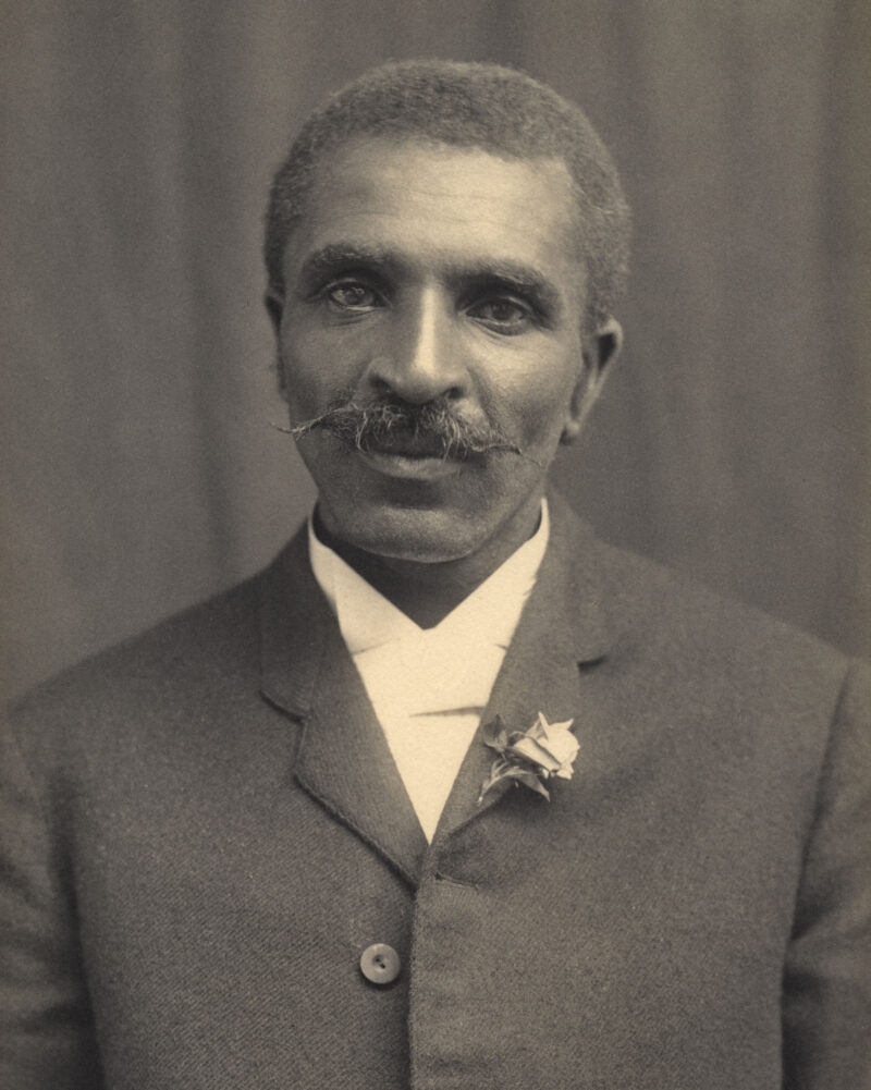 A black and white photo of an older man with a large moustache is seen from the waist up.