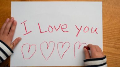 A child writing on printer paper with the words, "I Love You" in red crayons with hearts underneath